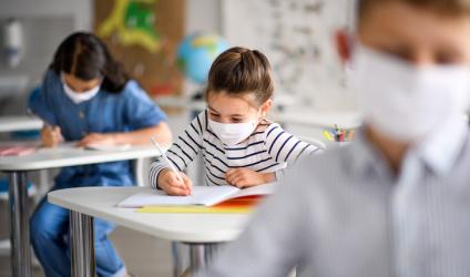 [image of 3 students in a classroom, seated at desks and writing. They are all wearing surgical masks]
