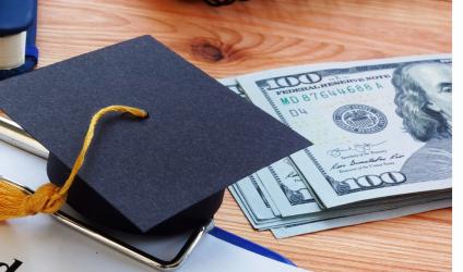 [image of a wooden table top with a small graduation cap decoration sitting next to a stack of $100 bills] 