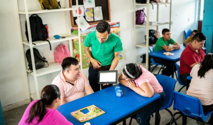 Photo of a teacher standing and showing a tablet to students who are sitting at a table 