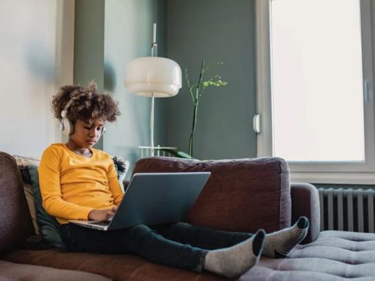 Young female African American student sitting on couch on her computer with headphones, focusing on schoolwork