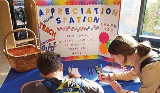 A male and a female student sit writing notes in front of a bulletin board reading "Appreciation Station"