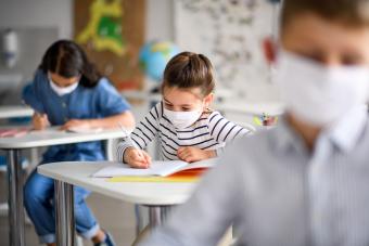 [image of 3 students in a classroom, seated at desks and writing. They are all wearing surgical masks]